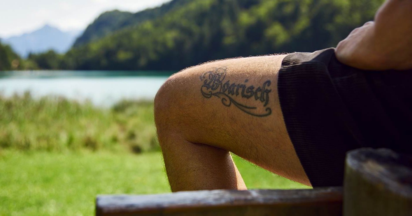 Schriftzug "Boarisch" tattowiert auf einem Oberschenkel, vor Landschaft